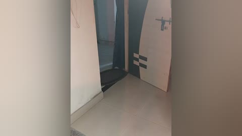 Media: Video of a narrow, dimly lit hallway with beige walls, a white door with black stripes, and a partially visible door to the left, leading to a bathroom with a white toilet. The floor is tiled in a light beige color.