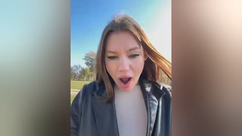 Media: Video of a young woman with light skin and brown hair, wearing a black jacket over a white top, outdoors on a sunny day, mouth open in an exaggerated expression.