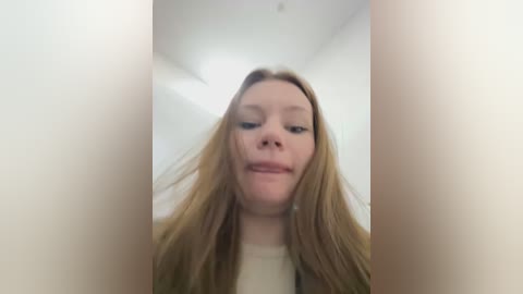 Media: A close-up video of a young Caucasian woman with long, straight blonde hair, fair skin, and a neutral expression. She is indoors, with a minimalistic white background and a slight blurriness.