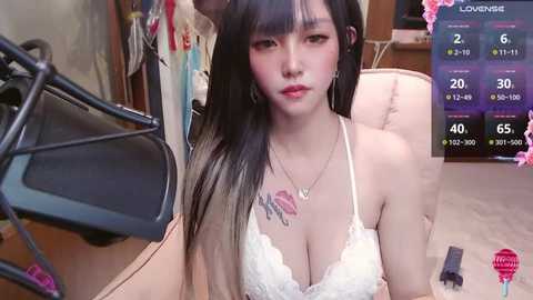 Media: Video of an Asian woman with long black hair, wearing a white lace bra, sitting in a beige chair, in a cluttered room with clothes hanging, a microphone, and a TV screen displaying streaming metrics.