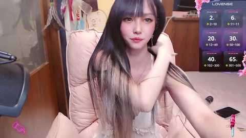 Media: Video of an Asian woman with long black hair, fair skin, and red lipstick, sitting on a beige chair, holding her hair, in a room with wooden furniture and a digital screen displaying a live stream.