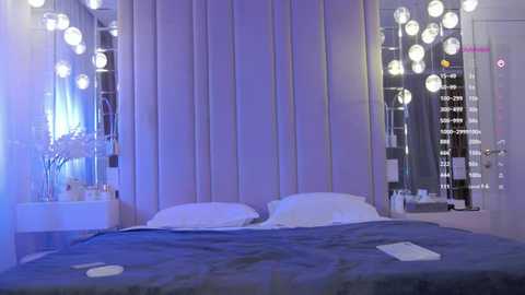 Media: Video of a modern bedroom with a plush, gray velvet headboard, white pillows, and a dark blue blanket. The walls are adorned with large, round, glowing white orbs.