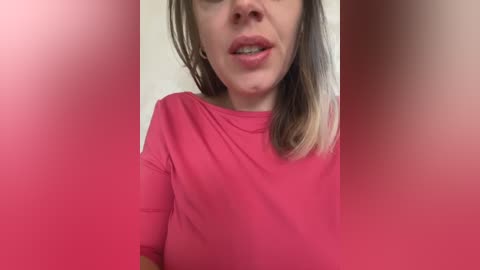 Media: Video of a fair-skinned woman with shoulder-length brown hair, wearing a pink shirt, mouth slightly open, taken in a pink-toned room.