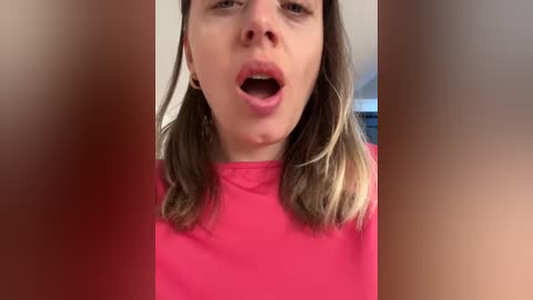 Media: Video of a young Caucasian woman with light skin and shoulder-length brown hair, wearing a bright pink shirt. Her mouth is open in a surprised or shocked expression. Background is blurred.