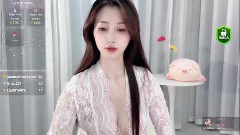 Media: Video of an East Asian woman with long black hair, wearing a white lace dress, seated in a modern room with soft lighting, pink and yellow flowers, and a white chair.