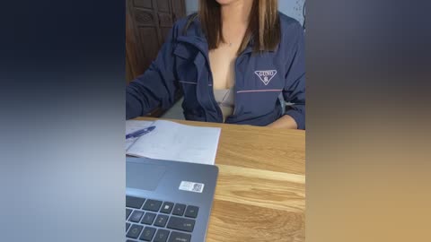 Media: A video of a woman with straight, shoulder-length brown hair, wearing a navy blue jumpsuit with a small triangle emblem on the chest, sitting at a wooden desk. A laptop and open book are on the desk.