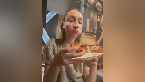 Media: Video of a blonde woman with closed eyes, puckering her lips, holding a burger with a white napkin in a modern restaurant.