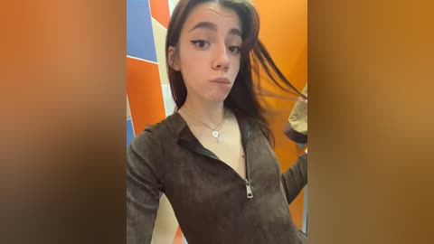 Media: Video of a young woman with light skin, dark hair, wearing a dark brown suede zip-up jacket, standing in a brightly colored room with orange and blue walls.