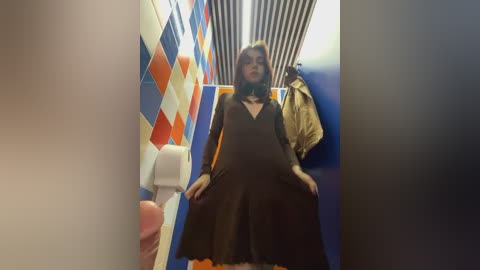 Media: Video of a woman with shoulder-length brown hair, wearing a long, black dress, standing in a public restroom with colorful, patterned tiles and a beige towel hanging on a wall.