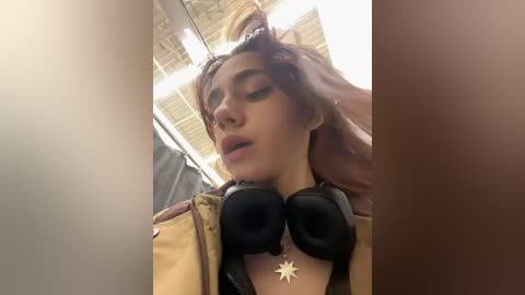 Media: Video of a young woman with long, wavy, reddish-brown hair, wearing a beige jacket, large black headphones, and a star necklace. Background shows a cloudy sky through a window.