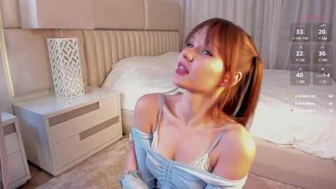 Media: Video of a young Asian woman with light skin and straight, shoulder-length auburn hair, wearing a light blue off-shoulder top. She sits on a beige carpet in a modern, minimalist bedroom with a white bed, nightstand, and white curtains.