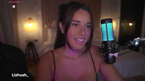 Media: Video of a young woman with medium brown skin, long dark hair in a ponytail, wearing a black spaghetti strap top, singing into a blue-lit microphone in a dimly-lit bedroom with a bed and nightstand.