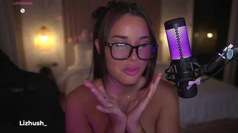 Media: Video of a young, topless woman with glasses and brown hair, wearing a microphone headset, recording in a dimly lit bedroom.