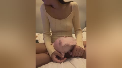 Media: Video of a woman in a cream-colored long-sleeve top, sitting on a white bed, cradling a pink plush toy against her bare chest, with her hands gently covering her breasts.