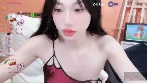 Media: Video of a young East Asian woman with fair skin, long black hair, and full lips, wearing a red satin camisole, in a cozy bedroom with a white bed, orange walls, and a TV showing an anime character.