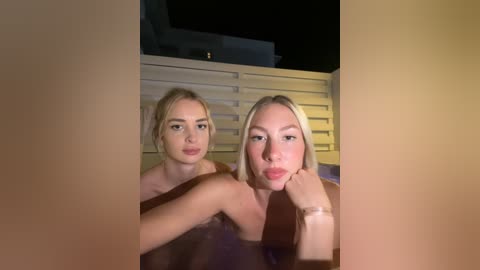 Media: Video of two blonde women in a hot tub, both topless, at night, with a modern house and fence in the background.