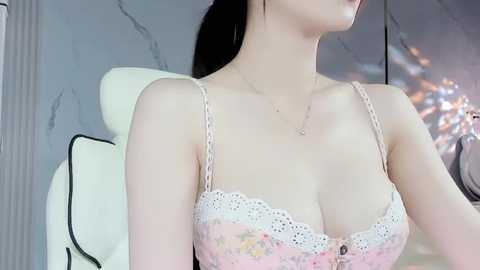 Media: Video of a slender Asian woman with fair skin and dark hair, wearing a pink floral lace bra with thin straps, indoors with a white chair and gray marble wall background.