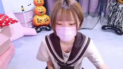 Media: Video of a young Asian girl in a school uniform, wearing a face mask, sitting in a Halloween-themed room with a pink bed, jack-o'-lanterns, and a black spider web.