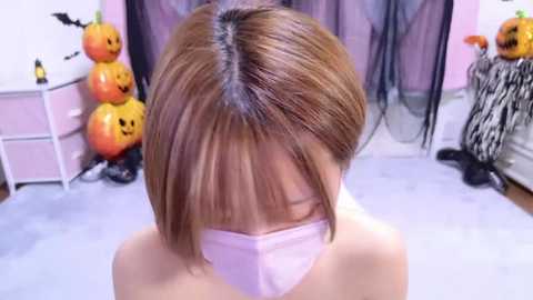 Media: Video of a young Asian woman with shoulder-length, straight brown hair, wearing a light pink mask, standing indoors. Background includes Halloween decorations, including carved pumpkins and black-and-white striped curtains.