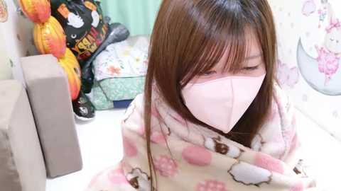 Media: A video of an East Asian woman with straight, shoulder-length brown hair, wearing a light pink face mask and a floral-patterned blanket, sitting on a couch with Japanese decor in the background.