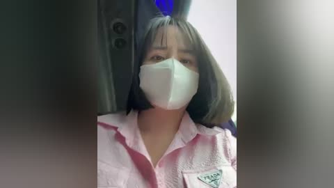 Media: Video of a woman wearing a pink button-up shirt and a white surgical mask, seated in a dimly lit room with a blue hat and a visible door handle in the background.