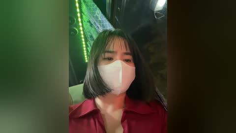 Media: A video of an Asian woman with shoulder-length black hair, wearing a white surgical mask and a red shirt, sitting in a dimly lit, possibly industrial or medical setting with green LED lights in the background.