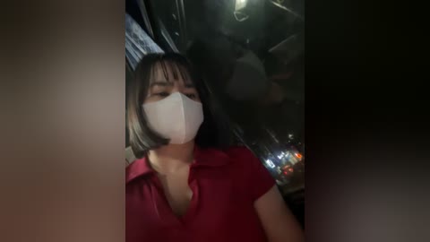 Media: Video of a young Asian woman with straight black hair, wearing a red shirt and white face mask, standing in a dimly lit, cluttered room with a fish tank in the background.