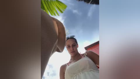 Media: A video featuring a young woman with a pale complexion, wearing a sheer, white dress, standing under a bright blue sky with clouds, and a palm tree in the background.