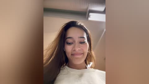 Media: Video of a young woman with medium-brown skin and straight, long brown hair. She has a slight smile and wears a white shirt. The background shows an indoor setting with beige walls and a ceiling fan.