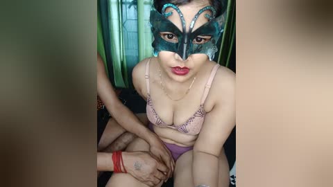 Media: Video of a woman with light brown skin, wearing a teal masquerade mask and pink lace bra, sitting on a bed, restrained by a man's red wristband around her wrist.