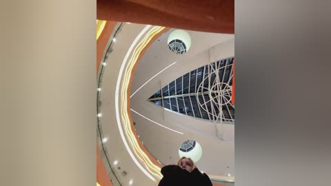 Media: Video of a modern, circular atrium with a central clock-like structure, featuring white walls, circular lights, and an orange railing. A person in a black coat stands in the foreground, slightly blurred.