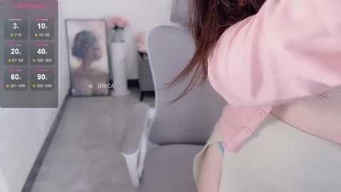 Media: Video of a woman in a pastel pink shirt standing in a modern, minimalist room with a gray chair, a white wall, and a digital screen displaying temperature data.