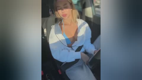 Media: A video of a blonde woman in a white hoodie and blue bra, driving, with sunlight streaming in.