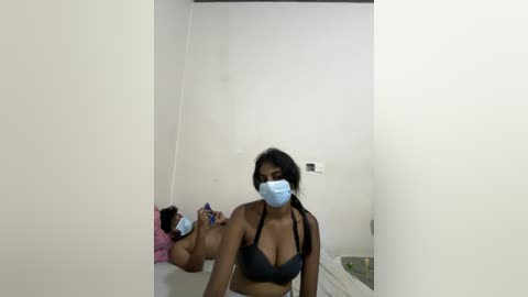 Media: Video of a woman in a black bra, wearing a blue surgical mask, lying on a bed with a pink blanket in a plain, white-walled room.