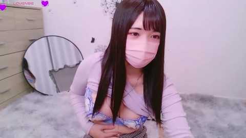 Media: Video of a young Asian woman with long black hair, wearing a light pink face mask and a revealing, low-cut top, lying on a plush carpet in a softly lit bedroom.