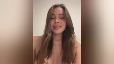 Media: A video of a young, light-skinned woman with long, wavy brown hair, smiling and topless, set against a plain beige background.