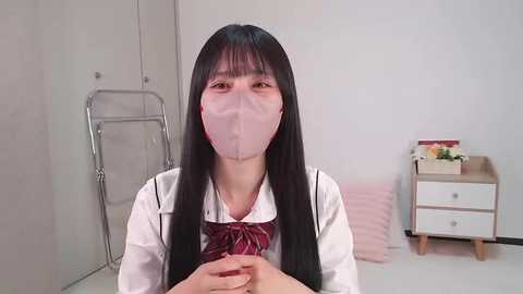 Media: Video of an Asian woman with long black hair and fair skin, wearing a white school uniform, pink mask, and red bow, standing in a minimalist, modern bedroom with light gray walls and a small wooden nightstand.