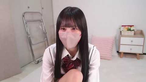 Media: Video of a young Asian woman with long black hair, wearing a white school uniform with a red tie, seated indoors. She has a surgical mask covering her nose and mouth. Background includes a metal folding chair, a small wooden desk, and a pink pillow.