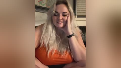 Media: Video of a smiling, fair-skinned woman with long, wavy blonde hair, wearing an orange tank top, resting her head on her hand in a cozy indoor setting.