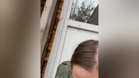 Media: Video of a person with light skin and short brown hair, wearing a green shirt, standing in front of a white window with a brown brick wall and a blurry tree outside.