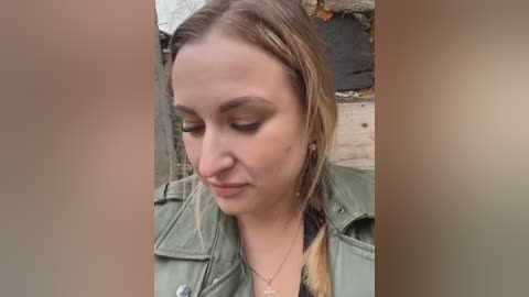 Media: Video of a young woman with fair skin, straight brown hair, and a green military-style jacket, sitting outdoors on a rustic wooden bench.