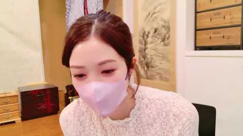 Media: Video of a young East Asian woman with pale skin and dark hair tied back, wearing a white lace blouse, pink surgical mask, and sitting at a desk. Background features a wooden chest, a black cabinet, and a white wall with a framed landscape painting.