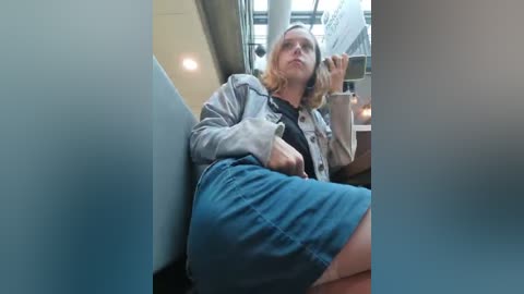 Media: Video of a young woman with light skin, shoulder-length blonde hair, wearing a gray hoodie and blue shorts, seated in a dimly lit train compartment, looking contemplative.