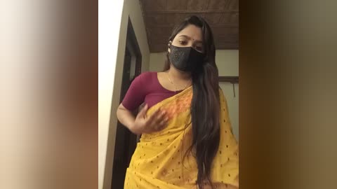 Media: Video of a woman with long dark hair, wearing a yellow sari with polka dots, a black face mask, and a maroon top. She stands in a dimly lit hallway with beige walls and a wooden ceiling.