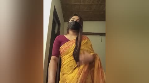 Media: Video of a woman in a maroon top, yellow sari, and black face mask, standing indoors with a wooden ceiling and beige walls.