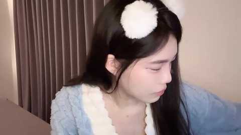 Media: Video of a young Asian woman with long black hair, wearing white panda headbands and a light blue fuzzy robe, sitting indoors against a brown curtain backdrop.