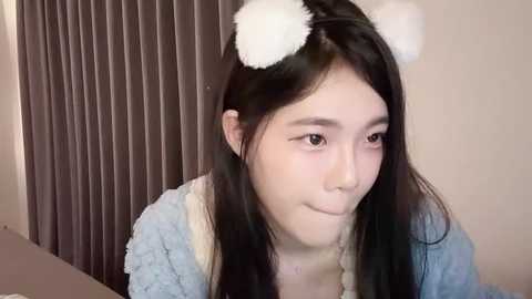 Media: A young Asian woman with long black hair, wearing white fuzzy bear ears and a light blue fuzzy cardigan, smiles softly in a cozy, dimly lit room with brown curtains.