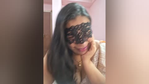 Media: Video of a woman with long black hair, wearing a black lace mask, lying on a bed with a beige blanket. She has a soft smile, with her hands resting on her face. The background shows a pink wall and a wooden door.