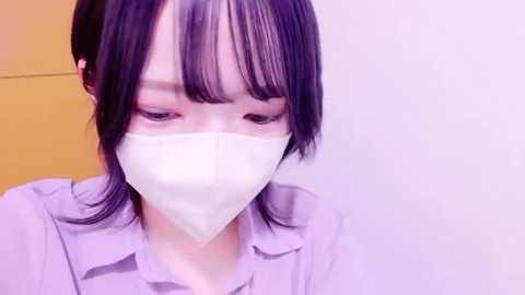 Media: Video of an Asian woman with pale skin, wearing a white surgical mask, black hair with bangs, in a light lavender shirt, against a plain white and yellow background.