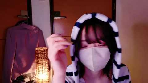 Media: Video of a woman with a white mask, wearing a striped hooded robe, holding a cigarette, standing in a dimly lit room with a wicker basket and hanging clothes in the background.